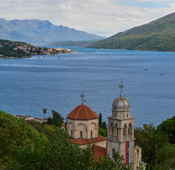 This landscape photograph captures a breathtaking view looking down on Savina Monastery, set against the stunning backdrop of Boka Bay in Herceg Novi, Montenegro. Taken during the late morning in early summer, the sunlight enhances the vivid blue of the bay and the lush green hills surrounding the water. The distinct red-tiled roofs and bell tower of the Savina Monastery stand out in the foreground, illustrating the rich architectural heritage of Montenegro. In the background, the expansive waters of Boka Bay stretch toward distant coastal towns and the impressive Orjen mountain range, creating a dramatic and peaceful scene unique to Herceg Novi.